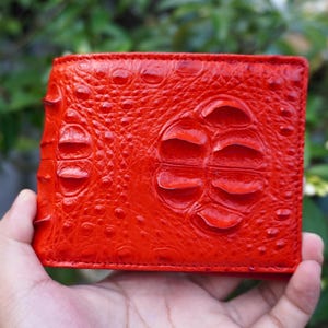 Può includere: Un portafoglio bifold rosso brillante caratterizzato da una texture in pelle di coccodrillo in rilievo. La superficie presenta squame grandi e arrotondate ben distinte e dettagli più piccoli e granulosi su tutto l'esterno, creando un aspetto tattile.