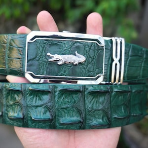 May include: A dark green belt with a textured, scale-like pattern resembling reptile skin. The belt features a rectangular silver-toned metal buckle with a polished finish and a small, detailed silver-toned alligator emblem centred on the buckle face.