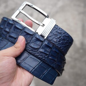 Op de afbeelding: Een donkerblauwe riem met een gestructureerd, reliëfpatroon dat het uiterlijk van reptielenschubben nabootst. De riem is voorzien van een gepolijste, rechthoekige zilverkleurige metalen gesp met een geribbeld detail aan de zijkant van het frame.