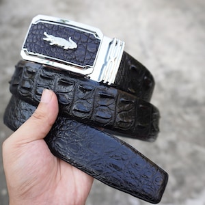 Puede incluir: Un cinturón marrón oscuro con un patrón texturizado en relieve que imita la piel de cocodrilo. El cinturón cuenta con una hebilla rectangular de metal plateado con acabado pulido y un pequeño emblema de cocodrilo metálico en el centro.