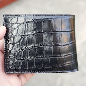 Peut inclure: Un portefeuille noir à deux volets présentant une finition brillante et un motif texturé en peau de crocodile. Cet accessoire rectangulaire affiche un design à écailles distinct sur sa surface, avec des coutures visibles sur les bords.