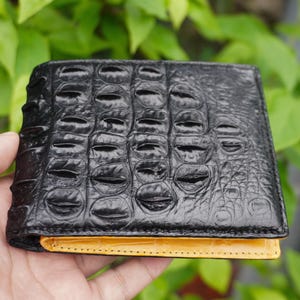 Könnte beinhalten: Ein schwarzes Bifold-Portemonnaie mit einer strukturierten Krokodilleder-Optik auf der Außenseite. Das Portemonnaie hat ein leuchtend gelbes Innenfutter, das an der Unterkante sichtbar ist, sowie sichtbare Nähte an der Lederkonstruktion.