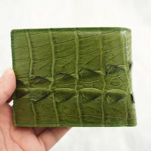 May include: A square-shaped bi-fold wallet featuring a textured olive green exterior with a distinct, raised scale pattern reminiscent of reptile skin. The wallet is constructed with visible green stitching along the edges, creating a uniform border.