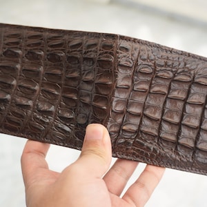 May include: A dark brown bifold wallet featuring a textured, raised scale pattern across the entire exterior surface. The wallet is rectangular in shape with visible stitching along the edges, showcasing a glossy finish and a distinct, repeating grid-like texture.