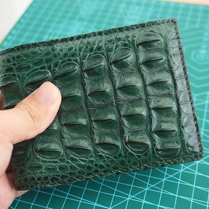 May include: A dark green bifold wallet featuring a textured, raised scale pattern across the exterior surface. The wallet has visible stitching along the edges and a glossy finish, showcasing a distinct, repeating organic texture throughout the design.