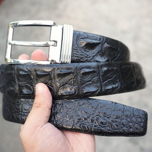 Può includere: Una cintura nera con un motivo a scaglie in rilievo e una fibbia in metallo argentato lucido. La cintura presenta una cornice rettangolare con ardiglione e un dettaglio a coste sul lato del meccanismo della fibbia, con una finitura lucida.