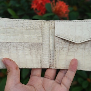 Könnte beinhalten: Ein aufklappbares Portemonnaie in Off-White mit einer strukturierten Krokodilleder-Optik. Das Innere verfügt über mehrere horizontale Kartenfächer auf der linken Seite und ein Münzfach mit dreieckigem Klappverschluss auf der rechten Seite.