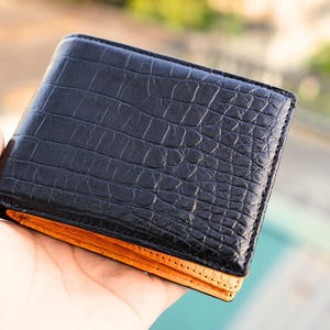 Puede incluir: Una cartera plegable con un exterior negro de textura de piel de cocodrilo y un forro interior de color naranja brillante que contrasta. La cartera tiene una forma rectangular con costuras visibles a lo largo de los bordes del cuero oscuro.
