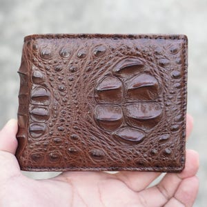 May include: A brown bifold wallet featuring a prominent, raised crocodile skin texture. The surface displays large, rounded, glossy scales in the centre, transitioning into smaller, intricate, pebbled scales towards the edges and the folded side.