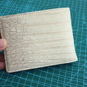 Può includere: Un portafoglio pieghevole con un esterno testurizzato color bianco sporco e un motivo in rilievo che imita la pelle di coccodrillo. Il portafoglio presenta cuciture visibili lungo i bordi e una forma rettangolare con una superficie dettagliata.
