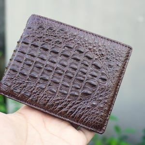 Può includere: Un portafoglio bifold marrone scuro caratterizzato da una texture in rilievo che imita la pelle di coccodrillo. La superficie esterna presenta un motivo ripetitivo di squame arrotondate e rialzate che riproduce la pelle di rettile.