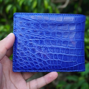 May include: A vibrant blue bifold wallet featuring a distinct, raised, and textured crocodile skin pattern across the entire exterior surface. The wallet has a rectangular shape with visible stitching along the edges, showcasing a glossy finish.