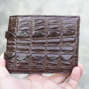 Può includere: Un portafoglio bifold marrone scuro caratterizzato da un motivo a squame in rilievo su tutta la superficie esterna. Il portafoglio ha una forma rettangolare con cuciture visibili lungo i bordi e una fila distinta di squame più grandi sul lato sinistro.