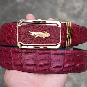 May include: A deep red belt with a textured, embossed pattern resembling reptile scales. The belt features a rectangular gold-toned metal buckle with a polished finish and a small, centred gold-toned alligator emblem on the front surface.
