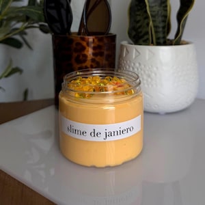 May include: A clear plastic jar filled with smooth, light orange slime topped with small yellow beads. A white label on the front of the jar features the black text "slime de janiero" in a serif font. The jar is positioned on a white surface.