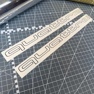 May include: Two white adhesive decals featuring the word quattro in a bold, black-outlined sans-serif font. The stickers are presented on a white backing paper, showcasing a clean, minimalist design suitable for application on smooth surfaces.