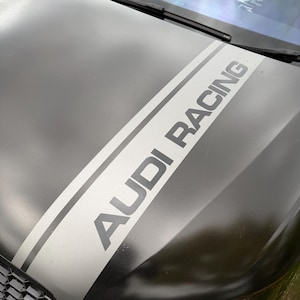 May include: A black car bonnet with a white and grey racing stripe running diagonally. The stripe displays the text "AUDI RACING" in grey. The car's headlights are visible at the bottom, and the windscreen wipers are at the top.