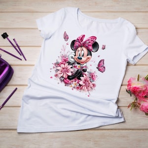 May include: White t-shirt featuring a Minnie Mouse graphic surrounded by pink flowers and butterflies. Minnie Mouse is wearing a pink bow and a pink and white outfit. The design is centered on the shirt.