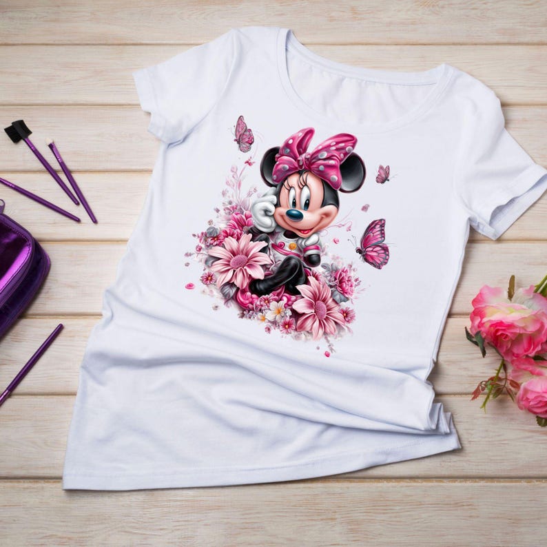 May include: White t-shirt featuring a Minnie Mouse graphic surrounded by pink flowers and butterflies. Minnie Mouse is wearing a pink bow and a pink and white outfit. The design is centered on the shirt.