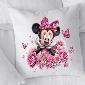 May include: A white pillow featuring a cartoon illustration of Minnie Mouse. Minnie Mouse is surrounded by pink flowers and butterflies. She is wearing a pink polka dot bow and dress. The pillow is on a white surface.