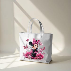 May include: White tote bag featuring a Minnie Mouse graphic. Minnie is wearing a pink dress and bow, surrounded by pink flowers and butterflies. The bag has two white handles and is made of a sturdy material.