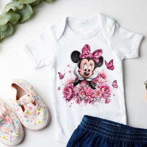 May include: White baby onesie featuring a Minnie Mouse graphic with a pink bow, surrounded by pink flowers and butterflies. The onesie is paired with a pair of pink and white baby shoes and a pair of blue denim shorts.