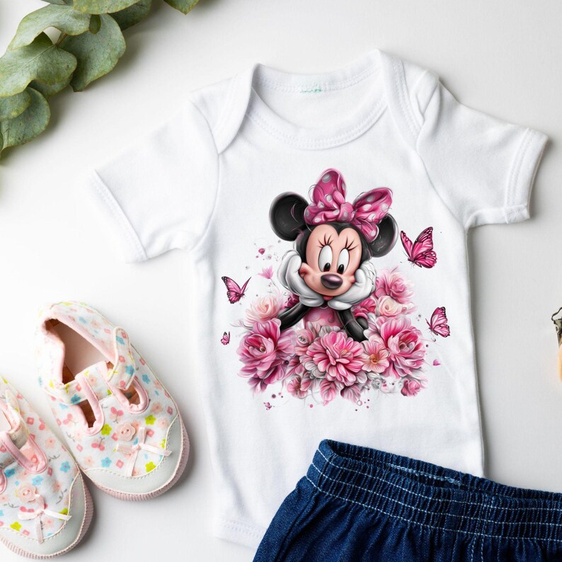 May include: White baby onesie featuring a Minnie Mouse graphic with a pink bow, surrounded by pink flowers and butterflies. The onesie is paired with a pair of pink and white baby shoes and a pair of blue denim shorts.