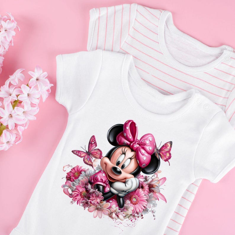 May include: White baby bodysuit featuring a Minnie Mouse graphic with a pink bow, surrounded by pink flowers and butterflies. A second white bodysuit with pink stripes is visible in the background. The background is a solid pink.