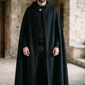 May include: A floor-length black hooded cloak featuring a single metallic circular clasp at the neckline. The garment is made of a solid dark fabric and drapes down to the ankles, worn over a black long-sleeved shirt and dark trousers.