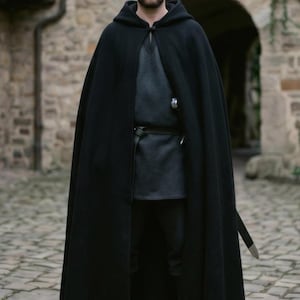 May include: A floor-length black hooded cloak made of heavy fabric, featuring a single button closure at the neck. The garment is worn over a dark grey tunic and black trousers, with a black belt and a sword scabbard visible at the waist.