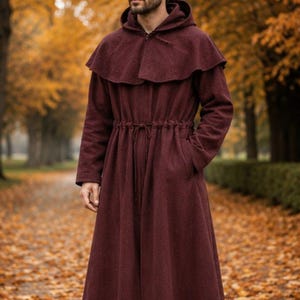 Op de afbeelding: Een enkellange, diepbordeauxrode mantel met capuchon, voorzien van een trekkoord in de taille, lange mouwen en een brede schoudercape. Het kledingstuk is gemaakt van een textuurrijke stof en wordt gedragen over een donkere broek met bruine leren laarzen.