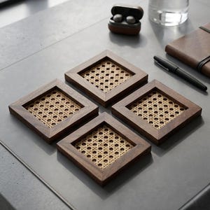 May include: A set of four square coasters featuring dark brown wooden frames and light beige woven rattan centres. Each coaster has a distinct grid pattern created by the natural cane webbing, providing a stable surface for drinks and glassware.