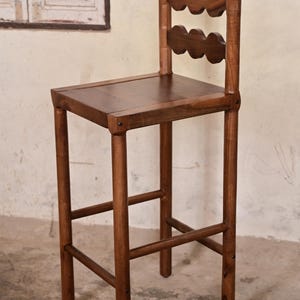 Puede incluir: Un taburete de bar alto de madera con acabado marrón oscuro. El taburete cuenta con un asiento cuadrado y un respaldo compuesto por dos listones horizontales con un borde festoneado en forma de nube. Tiene cuatro patas rectas unidas por travesaños.