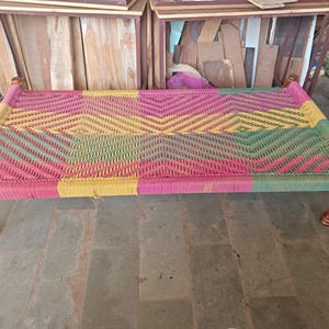 Könnte beinhalten: Ein gewebter Bettrahmen mit einem farbenfrohen geometrischen Muster. Das Bett hat ein lebendiges Design mit rosa, gelben und grünen Farbtönen. Der Rahmen ist aus Holz gefertigt und hat vier dekorative Beine.