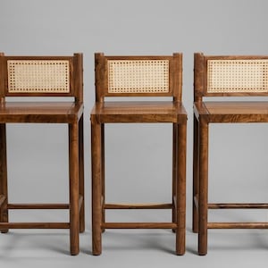 May include: Three tall bar stools constructed from dark brown wood, each featuring a square seat and a rectangular backrest with a light beige woven cane panel. The stools have four straight legs connected by horizontal support bars on all sides.