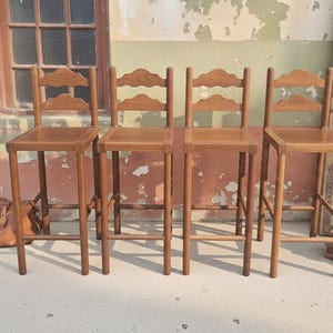 May include: A set of four tall wooden bar stools with square seats and straight legs. Each stool features a backrest with two horizontal slats, where the top slat has a decorative arched shape. The wood has a medium brown finish throughout.