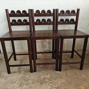 Puede incluir: Tres taburetes de bar de madera marrón oscuro con asientos cuadrados y cuatro patas rectas. Cada taburete cuenta con un respaldo compuesto por dos listones horizontales, decorados cada uno con una fila de cuatro motivos circulares tallados.