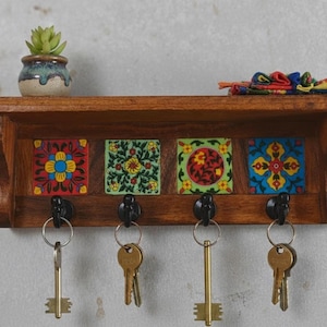 May include: A wooden key rack with four black hooks and decorative, colourful tiles. The shelf above holds a small potted succulent and a decorative fabric piece. Keys hang from the hooks, ready for use.