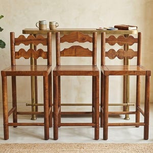 Könnte beinhalten: Drei Barhocker aus Holz mit mittelbraunem Finish, jeweils mit quadratischer Sitzfläche, vier geraden Beinen mit Fußstützen und einer Rückenlehne aus zwei horizontalen Lamellen mit markantem, wellenförmigem Randdesign.