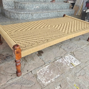 May include: A woven rope bed with a rectangular frame and four wooden legs. The bed's surface is made of tightly woven natural-coloured rope, creating a textured pattern. The wooden legs and frame have a warm, brown finish.