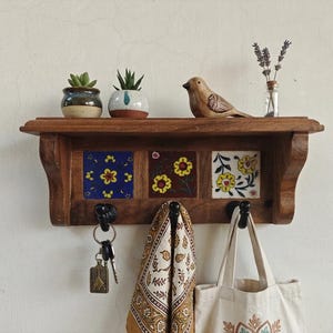 May include: A wooden wall shelf featuring three decorative ceramic tiles with floral patterns and three black metal hooks. The shelf holds two small potted succulents, a wooden bird figurine, and a glass bottle with dried lavender stems.