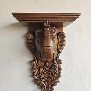 Puede incluir: Un estante de pared de madera marrón con una cabeza de elefante tallada a mano en el centro. El elefante tiene la trompa levantada y orejas detalladas. Debajo de la cabeza, el estante está decorado con intrincados patrones de hojas talladas.