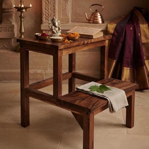 May include: A two-tier wooden step stool with a dark brown finish. The top tier holds a small silver-toned Ganesha figurine, three small metal bowls containing red powder, white grains, and orange flowers, and an open book with printed text.