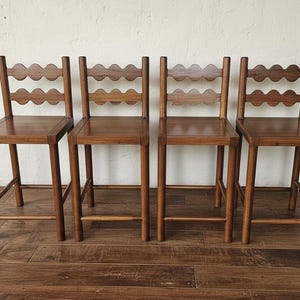 Puede incluir: Un juego de cuatro taburetes de bar de madera a juego con un acabado marrón medio. Cada taburete cuenta con un asiento cuadrado, cuatro patas rectas con travesaños de unión y un respaldo formado por dos listones horizontales con borde ondulado.