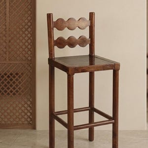 May include: A tall wooden bar stool with a dark brown finish. The stool features a square seat and a backrest composed of two horizontal rails, each with a scalloped, wavy edge design. Four straight legs are connected by a square footrest frame.