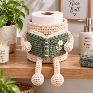 May include: A beige crochet toilet paper holder shaped like a character with arms and legs, holding an open dark green crochet book featuring a small white flower. The holder has a cylindrical body designed to fit a standard roll of toilet paper.