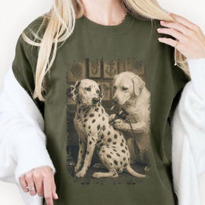May include: Olive green t-shirt featuring a sepia-toned illustration of a Dalmatian getting a tattoo from a Labrador. The Dalmatian has black spots, and the Labrador is holding a tattoo gun. The background includes tattoo flash art.
