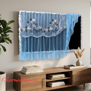 Op de afbeelding: Een lichtblauwe, doorschijnende televisiehoes met een geschulpte rand en geborduurde bloemmotieven in blauwe en witte tinten. De stof is transparant, valt over het scherm en heeft een decoratieve rand aan de boven- en zijkanten.
