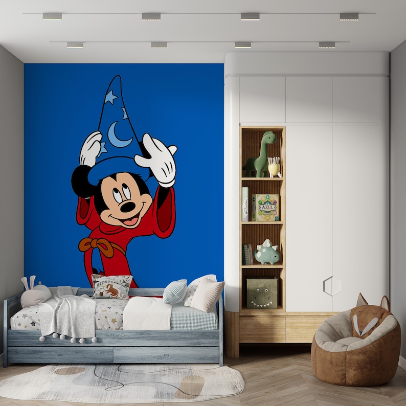 May include: A wall mural featuring Mickey Mouse as a sorcerer wearing a blue hat with moon and star patterns and a red robe, set against a solid blue background. The mural is positioned behind a bed with white bedding and a wooden storage unit.