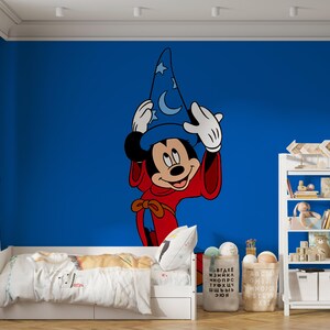 May include: A large wall decal featuring Mickey Mouse as a sorcerer, wearing a blue pointed hat with white moon and star patterns and a red robe. The character is positioned against a solid blue wall, centered above a white bed frame.